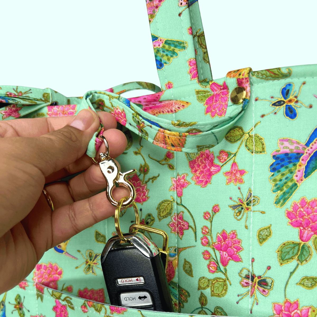 Close-up of a mint hummingbird, butterfly, and floral-print tote as a hand clips car keys to an exterior ring