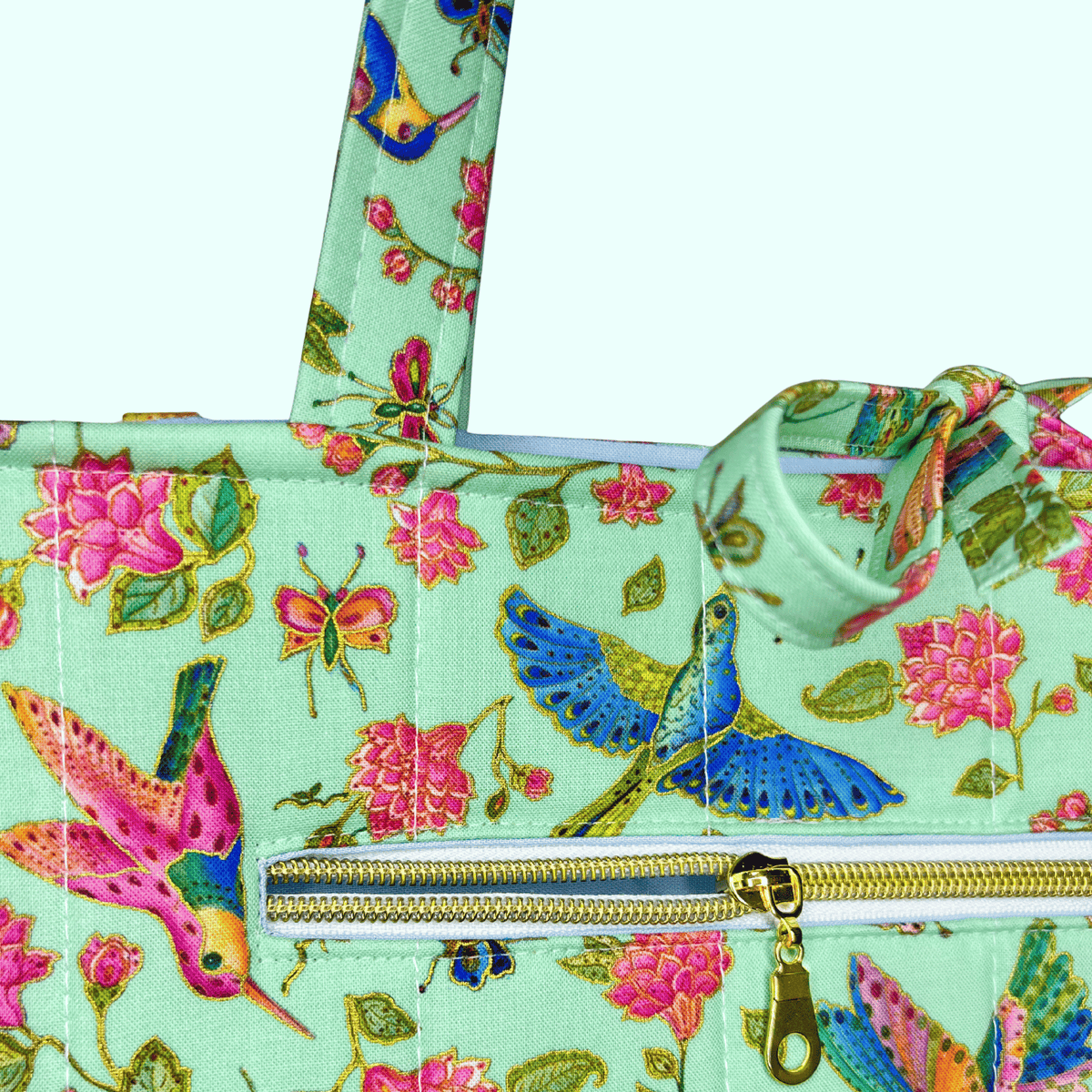 Close-up of a mint hummingbird, butterfly, and floral-print tote featuring a gold-zippered exterior pocket and tied handles.