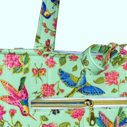 Close-up of a mint hummingbird, butterfly, and floral-print tote featuring a gold-zippered exterior pocket and tied handles.