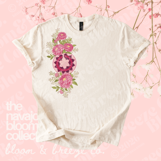 An off-white short-sleeve t-shirt with a colorful graphic print of pink flowers and a circular mandala design on the front, displayed on a light pink background with floral branch decorations.