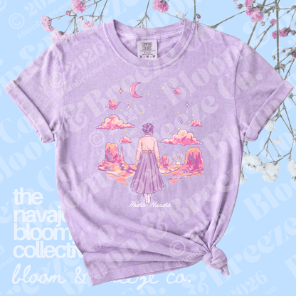 A light purple heather graphic t-shirt featuring an illustration of a girl in a pink dress looking at a sunset landscape with clouds and a crescent moon. The t-shirt is styled with a knot at the bottom hem and is displayed against a light blue background with decorative pink flowers.