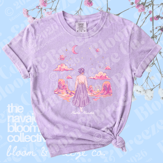A light purple heather graphic t-shirt featuring an illustration of a girl in a pink dress looking at a sunset landscape with clouds and a crescent moon. The t-shirt is styled with a knot at the bottom hem and is displayed against a light blue background with decorative pink flowers.