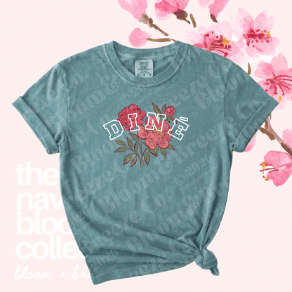 A teal short-sleeve t-shirt with a distressed finish, featuring a graphic of red and pink flowers with the word 'DINÉ' in white outlined letters. The shirt is styled with a knot at the bottom hem and is set against a light pink background decorated with cherry blossom branches.