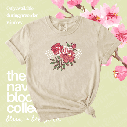 A beige short-sleeve t-shirt with a graphic of red and pink flowers, green leaves, and the word 'DINE' in white outline letters across the chest. The shirt is styled with a knot at the bottom hem against a light green background with pink cherry blossoms and text overlays.