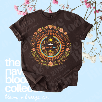 A dark brown short-sleeve t-shirt with a colorful circular graphic of the navajo Nation emblem, styled flat on a light blue background with decorative pink and white flowers.