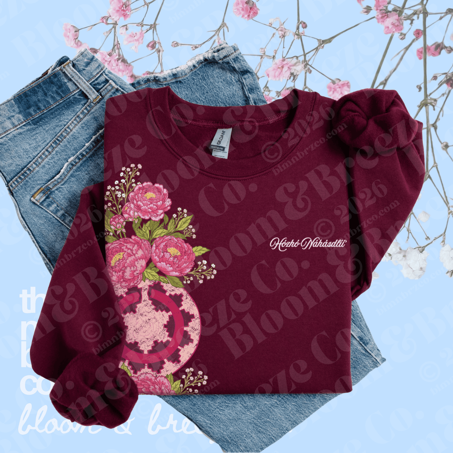 A folded long-sleeve crewneck sweatshirt in deep burgundy, featuring a large pink floral graphic on the front and a matching pink bow detail on the right shoulder. It is styled on top of blue jeans against a light blue background with pink blossoms.
