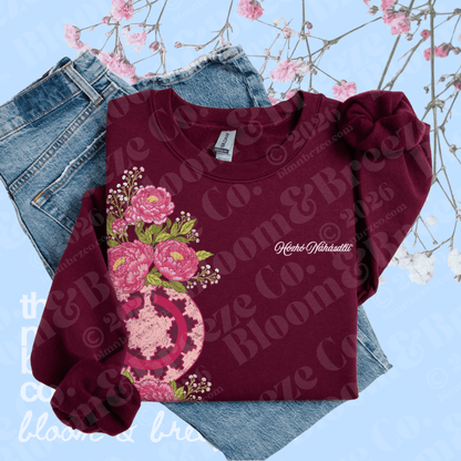 A folded long-sleeve crewneck sweatshirt in deep burgundy, featuring a large pink floral graphic on the front and a matching pink bow detail on the right shoulder. It is styled on top of blue jeans against a light blue background with pink blossoms.