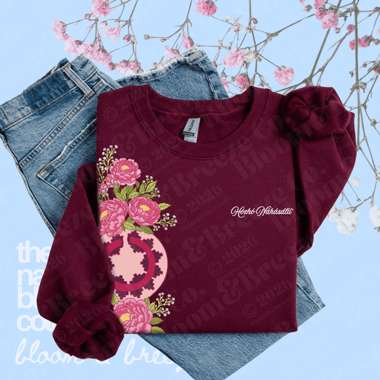 A folded burgundy long-sleeve shirt with a pink floral and peace-sign graphic on the front, styled over blue jeans with pink blossoms against a light blue background. The shirt has 'Katherine Martinez' printed on the chest.