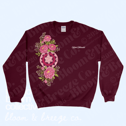A maroon crewneck sweatshirt featuring a large pink and green floral mandala graphic on the front, with 'Catherine Barnett' printed in white script on the left chest. The sweatshirt is displayed flat against a light blue background with a faint repeating watermark.