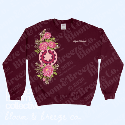 A crewneck sweatshirt in deep burgundy, featuring a large pink and green floral mandala graphic on the front and a small white script logo on the left chest. The sweatshirt is displayed flat against a light blue background with a repeating watermark pattern.