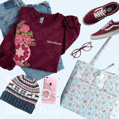 A styled flat-lay outfit arrangement featuring a burgundy long-sleeve top with a pink floral graphic and 'Amanda Michelle' text, paired with blue jeans, burgundy Vans sneakers, a light blue floral tote bag, a navy knit beanie, and a pink smartphone on a pale blue background.