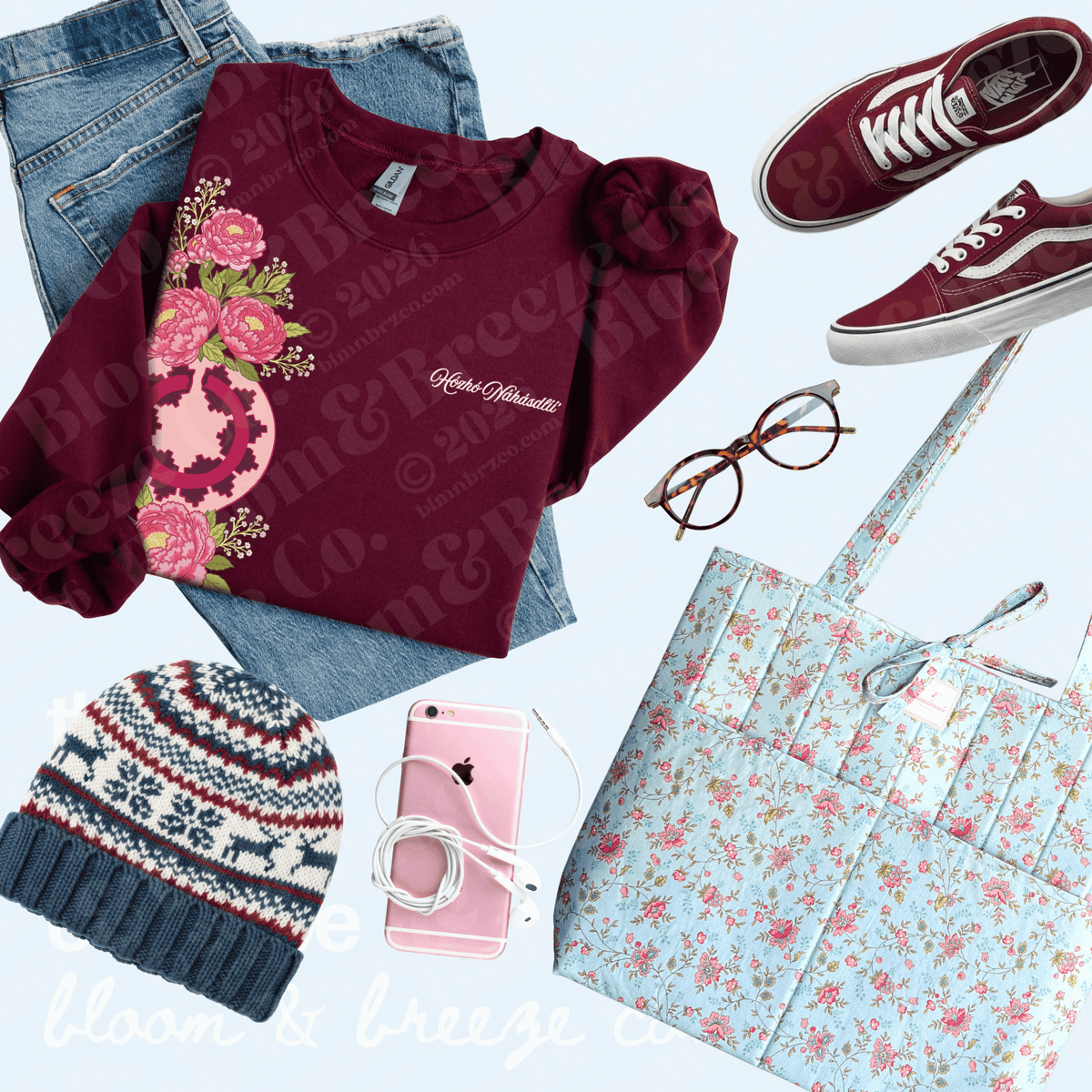 A styled flat-lay outfit arrangement on a light blue background featuring a burgundy long-sleeve top with a pink floral graphic and white script text, blue jeans, burgundy low-top sneakers, a light blue floral tote bag, a navy knit beanie, and eyeglasses.