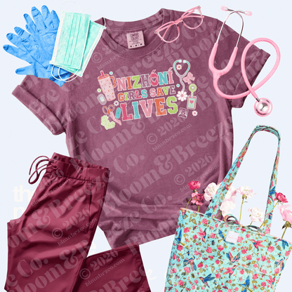 A coordinated medical-themed outfit set laid out on a light gray background, featuring a mauve short-sleeve T-shirt with a colorful nurse graphic reading 'NIZHONI GIRLS SAVE LIVES,' matching mauve scrub pants, blue gloves, a mint face mask, pink eyeglasses, a pink stethoscope, and a floral print scrub bag with flowers.