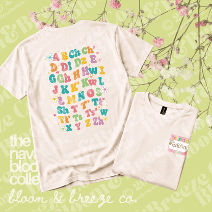 A white short-sleeve T-shirt shown from the back, featuring colorful alphabet letters with small flower graphics. A folded matching T-shirt is placed in front on a light green floral background.