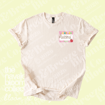 A light cream-colored short-sleeve t-shirt with a colorful graphic on the left chest that reads 'hello I’m the teacher' in navajo with a small pink apple logo, displayed on a pale yellow background with faint watermarks.