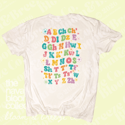 A white short-sleeve t-shirt with a colorful alphabet graphic on the front, displayed against a pale yellow-green background with faint watermarks.