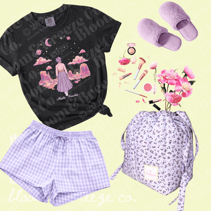 A flat-lay outfit set featuring a black graphic t-shirt with a pastel fantasy illustration, lavender gingham shorts, lavender slip-on slippers, and a matching lavender floral backpack, arranged on a pale yellow background with cosmetic props.