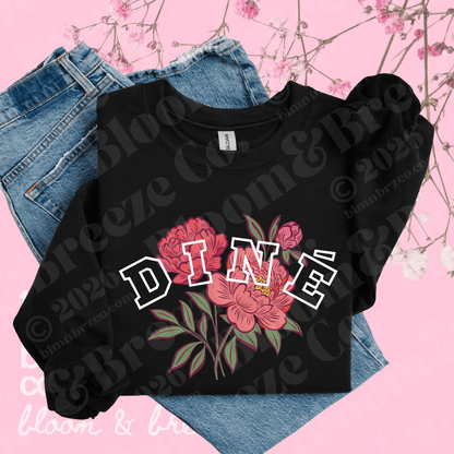 A black crewneck sweatshirt with a 'DINE' floral graphic, styled on top of blue jeans against a pink background with cherry blossom branches.