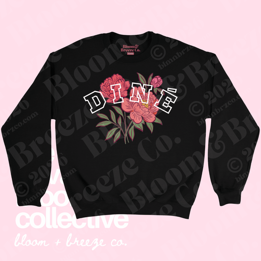 A black crewneck sweatshirt featuring a graphic print of pink peonies with green leaves and the word 'DINÉ' in white outlined letters across the chest. The sweatshirt is displayed flat against a light pink background with watermarks.