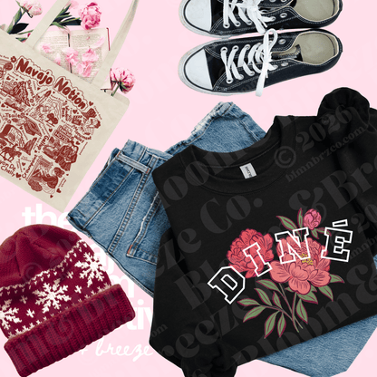A styled flat-lay featuring a black crewneck t-shirt with a pink floral graphic and the word 'DINE' in white outlined letters, layered over light blue denim shorts. The outfit is arranged on a pink background alongside a beige canvas tote bag, a burgundy knit beanie with white snowflake patterns, and a pair of black canvas sneakers.