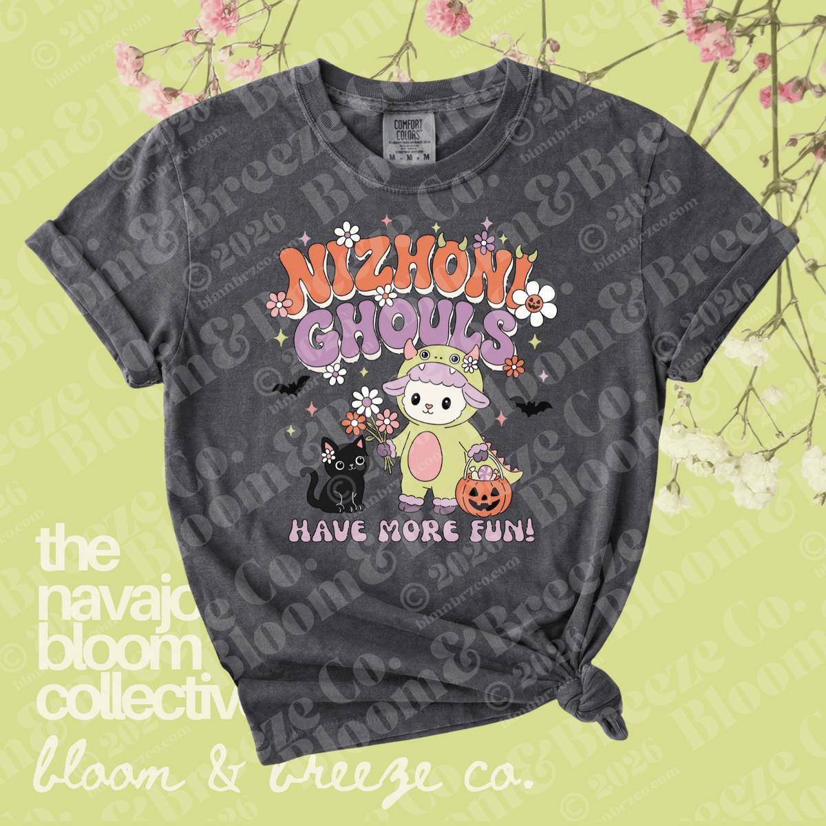 A dark gray graphic t-shirt with a distressed wash, featuring a cute cartoon ghost character in a pink bunny suit with the text 'NIZHONI GHOULS HAVE MORE FUN!'. The t-shirt is displayed flat with a knot at the bottom right, set against a light yellow-green background decorated with pink cherry blossoms and watermarked with 'the navajo bloom collective' and 'bloom & breeze co.