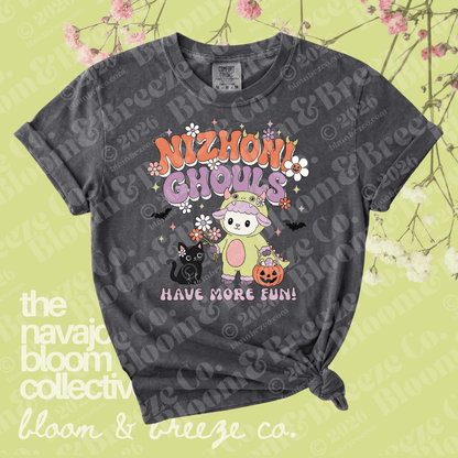 A dark gray graphic t-shirt with a distressed wash, featuring a cute cartoon ghost character in a pink bunny suit with the text 'NIZHONI GHOULS HAVE MORE FUN!'. The t-shirt is displayed flat with a knot at the bottom right, set against a light yellow-green background decorated with pink cherry blossoms and watermarked with 'the navajo bloom collective' and 'bloom & breeze co.