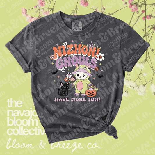 A dark gray graphic t-shirt with a distressed wash, featuring a cute cartoon ghost character in a pink bunny suit with the text 'NIZHONI GHOULS HAVE MORE FUN!'. The t-shirt is displayed flat with a knot at the bottom right, set against a light yellow-green background decorated with pink cherry blossoms and watermarked with 'the navajo bloom collective' and 'bloom & breeze co.