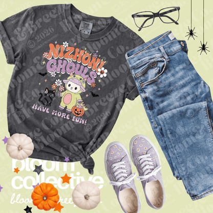 A styled flat-lay featuring a dark gray graphic t-shirt with 'Nizhoni Ghouls Have More Fun!' text and a cartoon cat in a Halloween costume. The shirt is styled with blue jeans, lavender sneakers, glasses, and small pumpkins on a pale yellow background with a 'bloom collective' watermark.