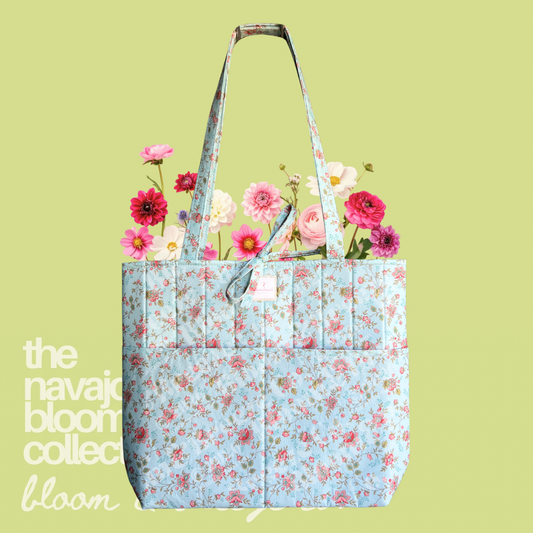 Light blue floral quilted tote bag with pink flowers, long straps, and front pockets on a soft green background.