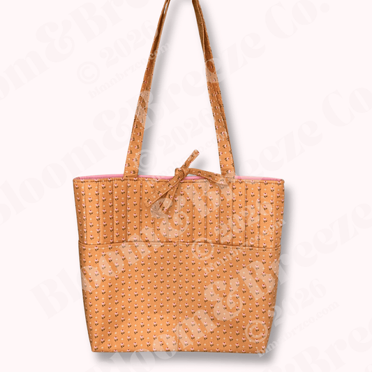 Quilted tote bag in warm honey mustard fabric with small pink and cream floral print, vertical stitching, long shoulder straps, and a front pocket detail.