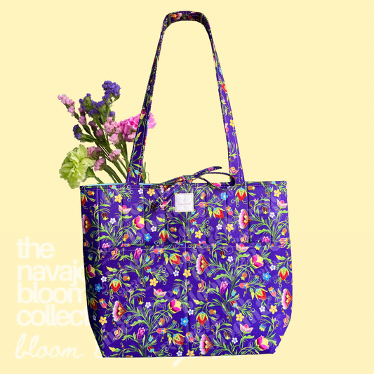 Purple floral quilted tote bag with long straps, front pockets, and flowers on a soft yellow background.