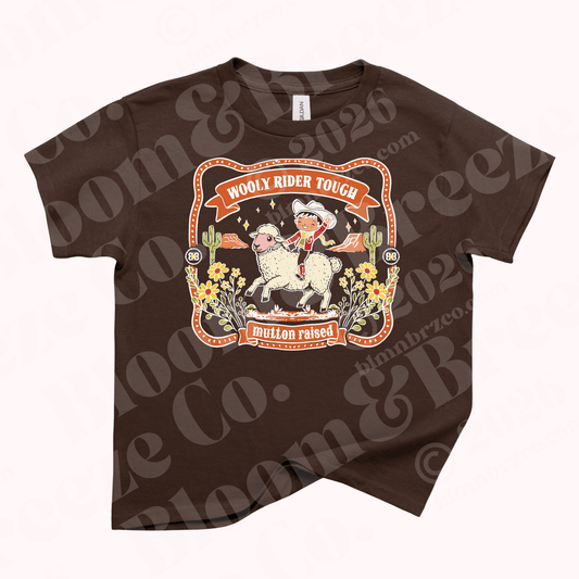 Brown youth t-shirt with vintage western style graphic of a child riding a sheep surrounded by desert flowers and cacti, reading “Wooly Rider Tough” and “mutton raised.”