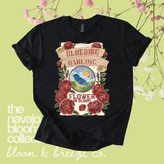 Black Bluebird Darling graphic tee with red flowers, a bluebird illustration, and “Flower in Bloom” text on a soft green floral background.