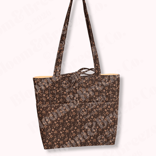 Quilted tote bag in rich brown fabric with delicate tan floral print, vertical stitched detailing, long straps, and a front pocket design.