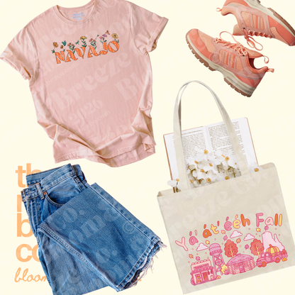 A styled flat-lay outfit featuring a light pink graphic t-shirt with 'NAVAjo' and floral details, folded blue denim shorts, coral pink sneakers, and a cream canvas tote bag printed with 'Y'all, beech fall' in pink and orange.