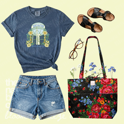 Blue Diné Nation water tower graphic tee styled with denim shorts, black sandals, glasses, and a black floral quilted tote bag.