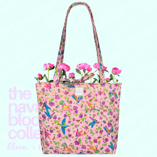 A pink fabric tote bag with a colorful floral and bird print, featuring long shoulder straps and pink flowers inserted at the top, set against a pale aqua background with faint pink text on the left.