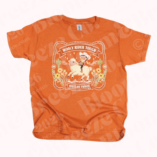 Orange t-shirt with a little cowboy riding bucking sheep and text reading wooly rider tough, mutton raised, buckle style graphic on a white background