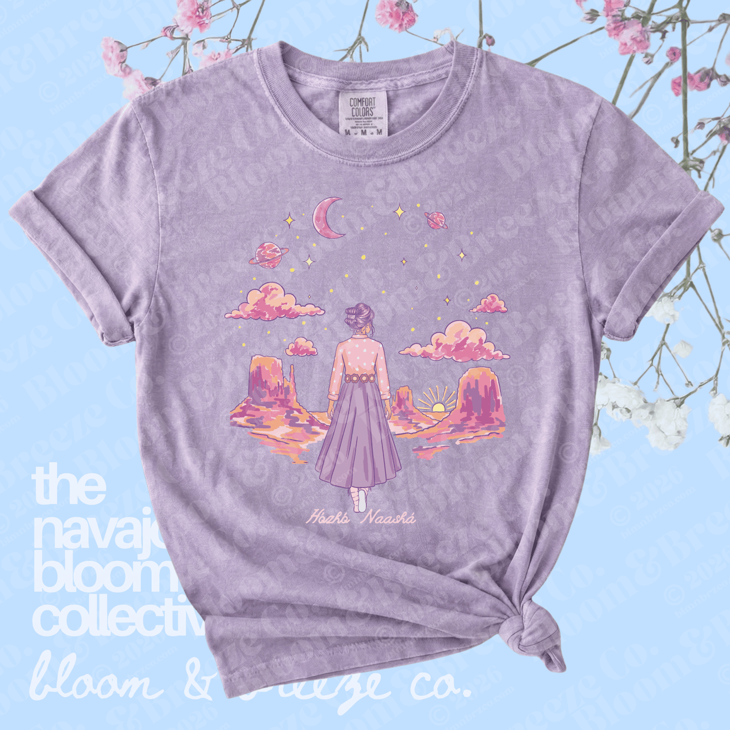 Purple t-shirt with a graphic design of a person in a desert landscape, featuring clouds and a moon, on a light blue background with floral elements.