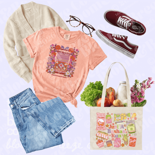 A styled flat-lay outfit featuring a coral pink graphic t-shirt, a cream cardigan, blue jeans, burgundy Vans sneakers, a canvas tote bag with colorful illustrations and groceries, and glasses, all arranged on a light lavender background.